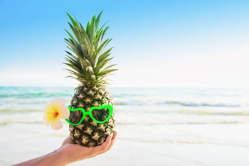 Hand holding a pineapple wearing heart sunglasses on a beach
