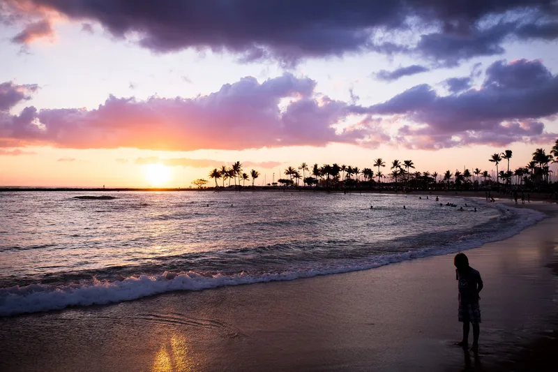 Person silhouetted on a Hawaiʻi beach at sunset with palm trees on the point