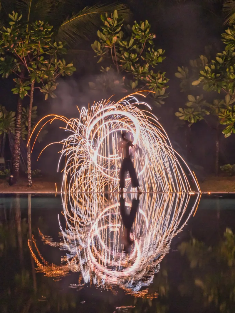 Fire spinner reflected in water among palm trees at night
