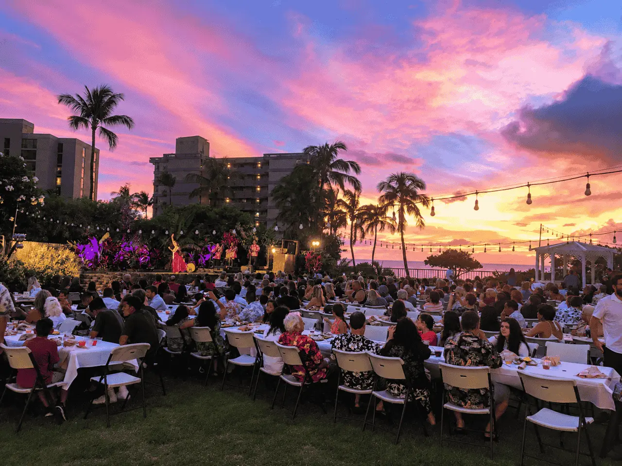 Guests enjoying an oceanfront Maui luau at sunset with vibrant pink and orange skies